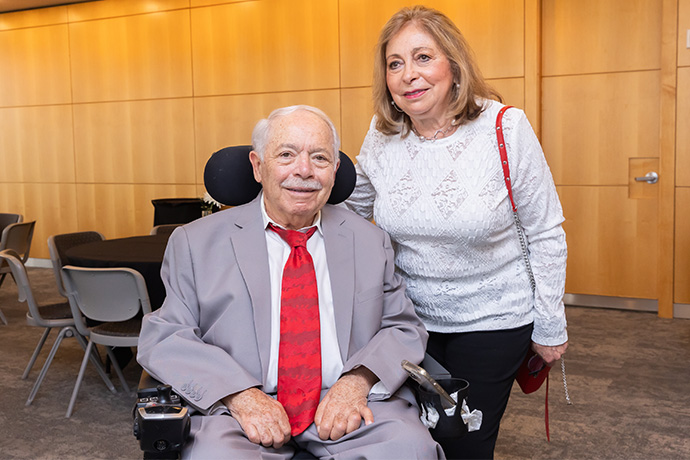 Smiling woman wearing a white blouse, in a conference room standing, next to a smiling gray-haired man wearing a gray suit and red tie, seated in a motorized wheelchair.