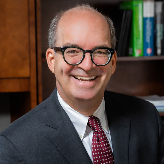 Smiling balding man, wearing a dark suit, white shirt, red tie, and dark-rimmed glasses, posed in front of a book case.