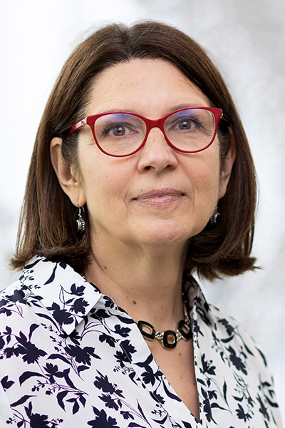 Smiling woman with dark hair, wearing a blouse with a dark flower print on a white background and red-rimmed glasses.
