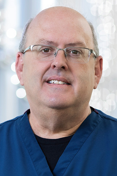 Smiling bald man, wearing blue scrubs with the UT Southwestern Medical Center logo and metal-rimmed glasses.