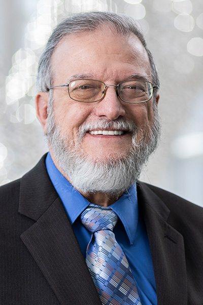 Smiling man with gray hair, beard, and mustache, wearing metal framed glasses and a dark suit with a blue shirt and tie.