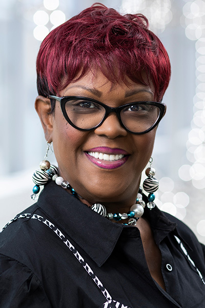 Smiling woman with red hair wearing a black dress with a black & white checked jumper and dark horn-rimmed glasses.