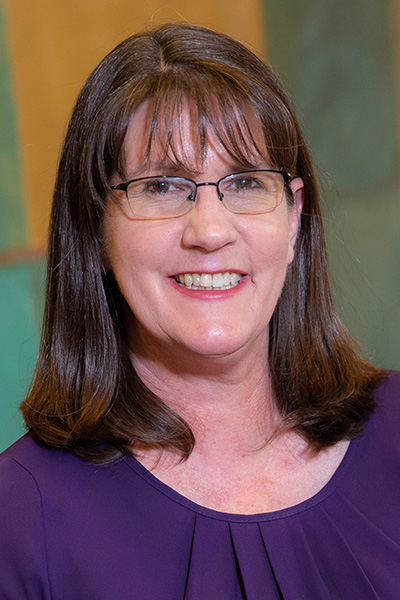 Smiling woman with shoulder lenth brown hair and bangs, wearing a purple blouse and dark rimmed glasses.