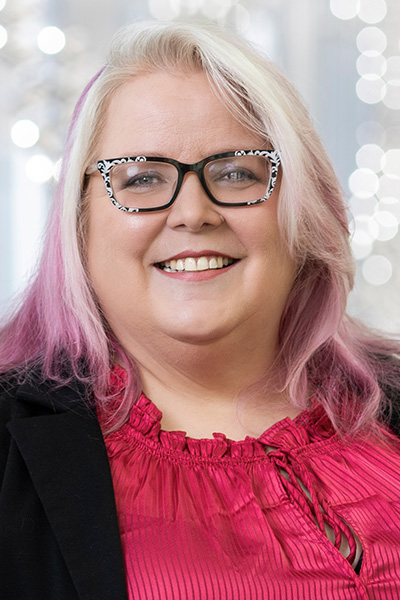 Smiling woman with purple and white hair, wearing a black jacket over a chartruese blouse and black and white print glasses.