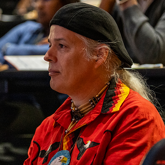 man in audience with beaded necklace and red shirt with eagle motif wears hat over long silver hair in ponytail