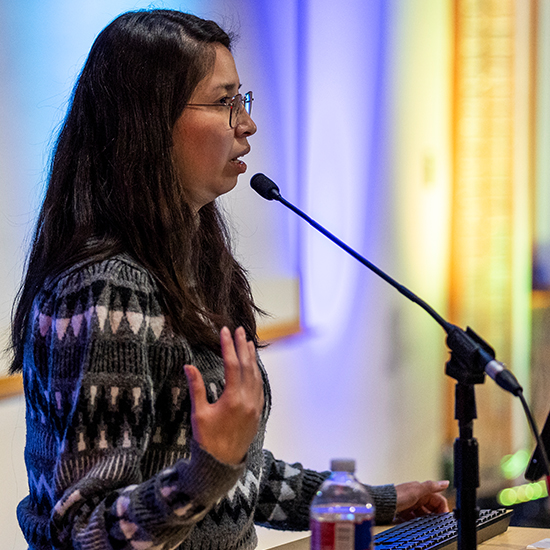 speaker at a podium with microphone is a woman with long black hair and glasses wearing a gray and white patterned sweater