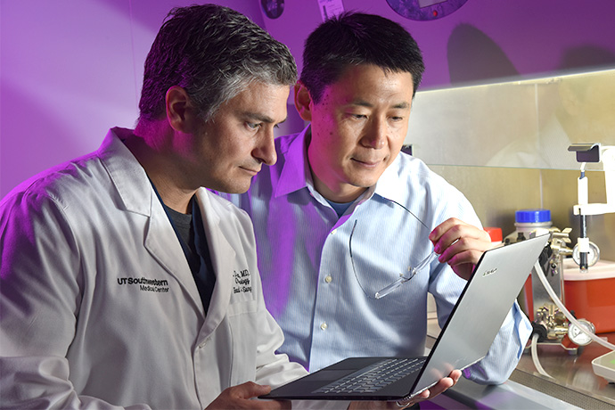 Dr. Sumer with grey hair wearing lab coat holds a laptop as Dr. Gao in blue shirt holding eyeglasses looks over his shoulder