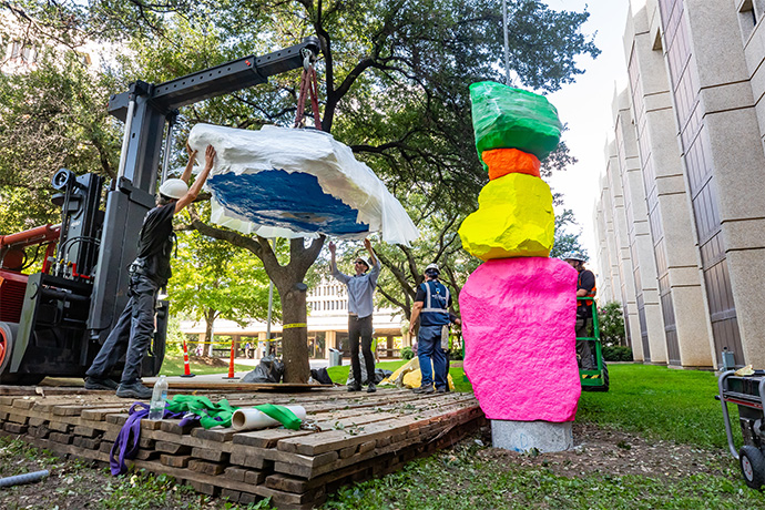 Small green crane hoisting a large blue boulder partially wrapped in plastic, poised to drop onto a tall gray pole, atop 5 other brightly colored boulders.