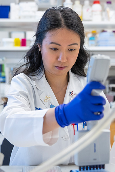 asian woman researcher with long dark hair wears blue gloves and white lab coat while watching results from a device in her hand