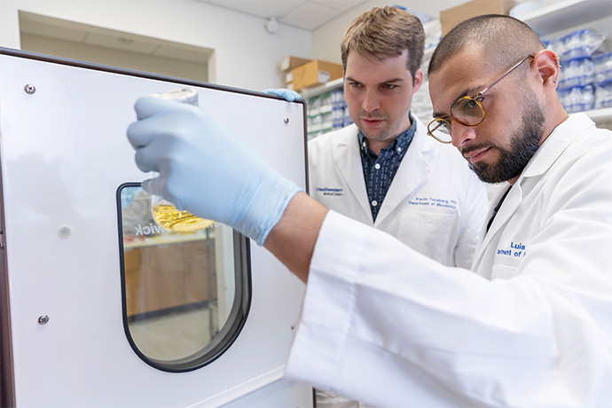 Two men with dark hair, wearing lab coats, in a lab, examining the yellow liquid content of a clear glass beeker.