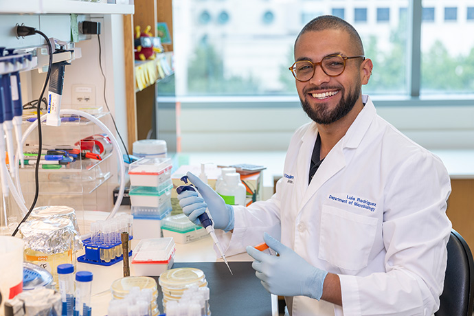 Smiing man with dark trim hair, beard, and mustache, wearing a white lab coat and brown framed glasses, sits at a cluttered lab desk hoding a beeker and dropper. Lab coat says - Luis Rodriguez, Department of Microbiology