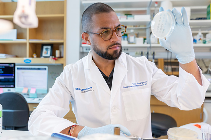 Man with trim dark hair, beard and mustache, in a lab, holding up a petrie dish to examine the content.