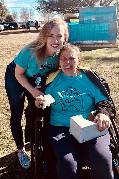 Smiling young woman with long blond hair, wearing a blue t-shirt, jeans, and tennis shoes, bending over posing with a blond woman wearing a blue t-shirt and jeans, seated in a wheelchair holding a box of tissue.