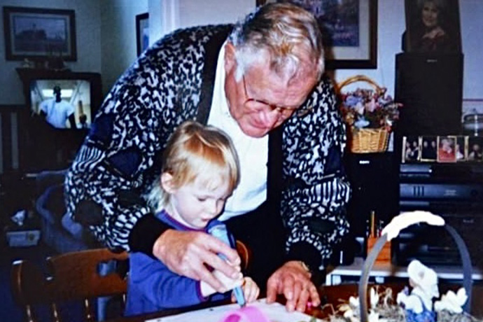 Man with gray hair, wearin a navy blue and white patterned sweater and glasses, bends over a young blond child seated at a dining table, helping them to write with a large pen.