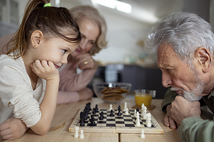 Blond woman watches as young girl and old man bend over a chessboard.