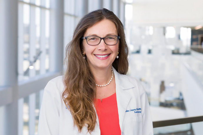 Smiling woman with long brown hair, wearing a white UT Southwestern Medical Center lab coat and dark-rimmed glasses.