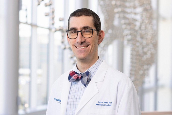 Smiling man with short dark hair, wearing a white UT Southwestern Medical Center lab coat, dark-rimmed glasses, and a bow-tie.