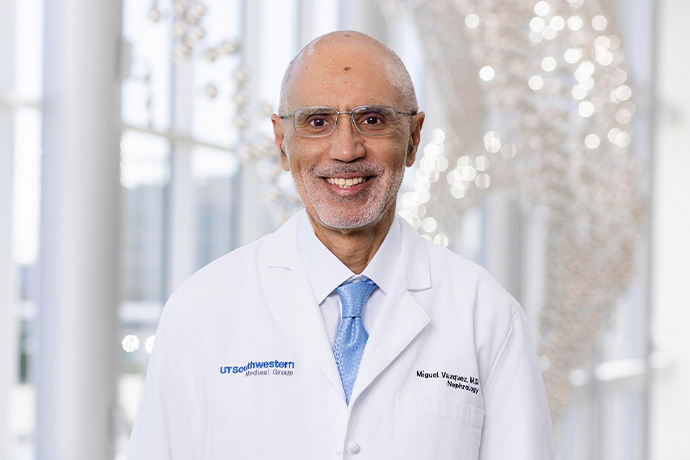 Smiling man with short-cropped white hair and facial scruff, wearing a white UT Southwestern Medical Center lab coat, and wire-rimmed glasses.