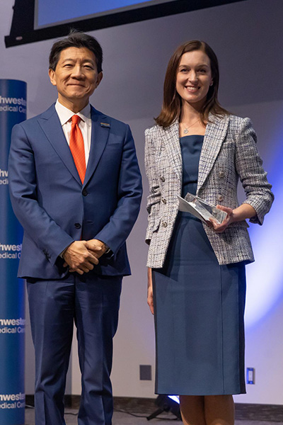 Smiling man with dark hair, wearing a blue suit and red tie poses with a woman with long brown hair, wearing a blue plaid jacket over a blue dress.