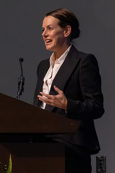 Woman with dark hair in a bun, wering a pin striped suit jacket and white blouse speaks from a podium.