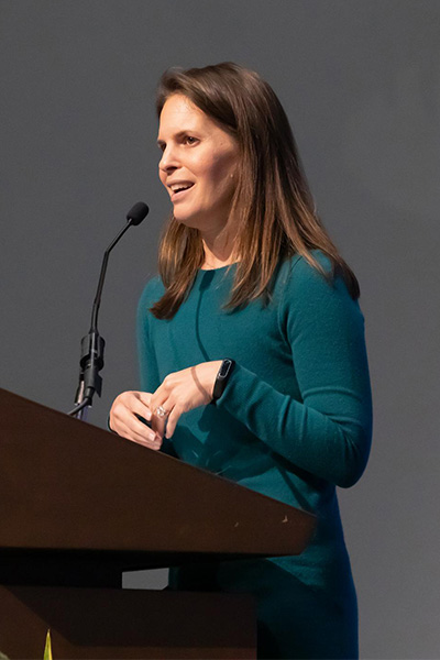 Woman with long brown hair, wearing a green sweater-dress speaks from a podium.