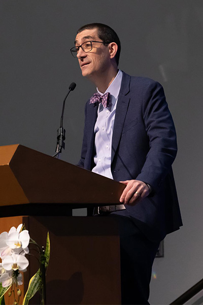 Man wearing a dark suit, purple shirt, purple bow-tie and dark rimmed glasses speaks from a podium.