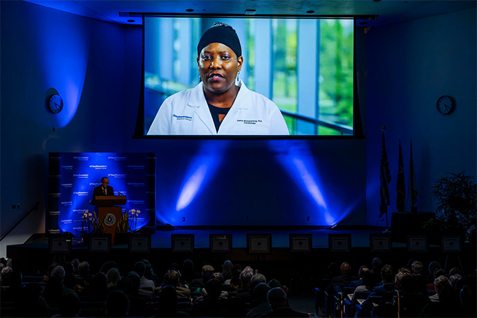 Woman wearing a dark cap and a white lab coat speaks on screen to large audience and man standing on stage at a podium.