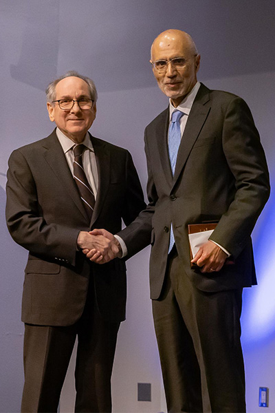 Two men men wearing dark suits and glasses, shake hands.