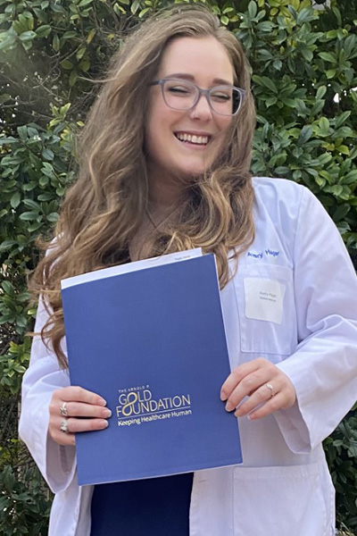 Smiling woman with long brown hair and glasses, standing in a courtyard wearing a lab coat over a horizontal striped dress, holding a blue notebook.