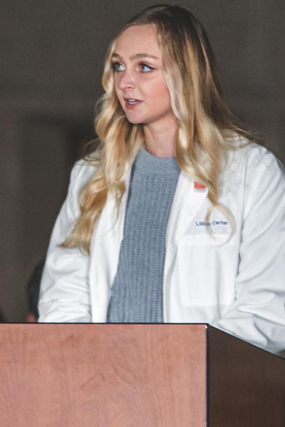 Woman with long blond hair, wearing a white lab coat, standing at a podium.