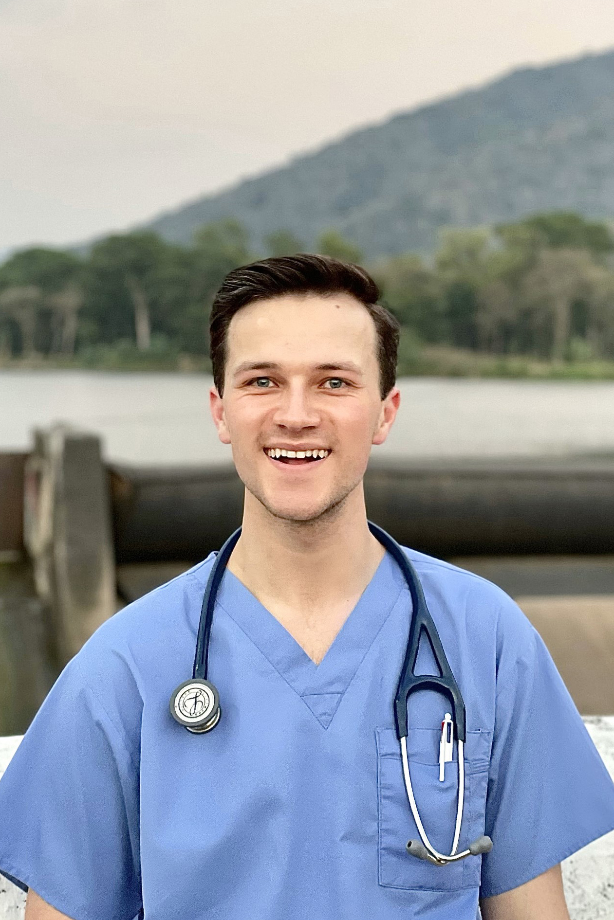 Smiling man with short, dark hair, wearing scrubs and a stethoscope, with lake and mountains in the background.