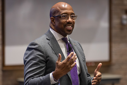 bald black man with glasses in brown suit and purple tie speaks on stage