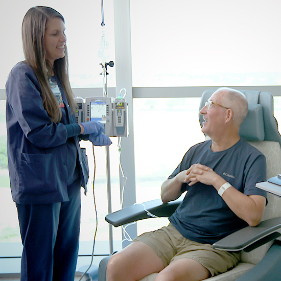 oncology nurse wearing blue scrubs stands talking to older male patient sitting in chemotherapy chair