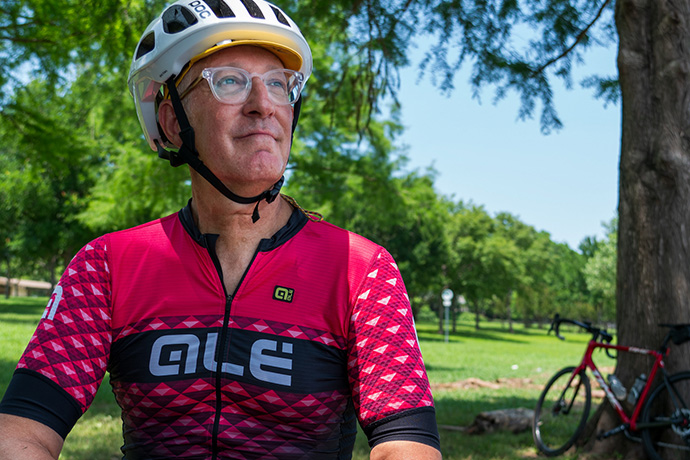 older man wearing jersey and cycling helmet stands near his bike and a tree outdoors
