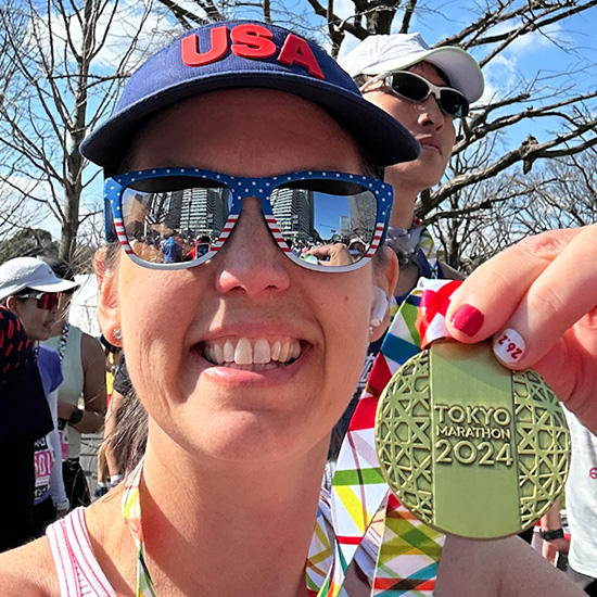 oncology nurse in hat and sunglasses holds up ribbon and medal from Tokyo marathon