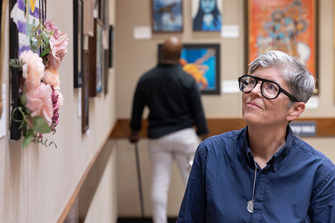 woman with short grey hair, glasses wearing blue shirt admires artwork hanging on a wall; black man with cane looks at art hanging in background