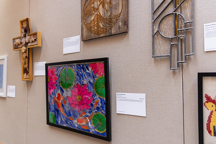 5 pieces of art hang on a beige wall: from left, a multimedia piece in the shape of a cross, pink water lilies in a frame, metal and glass sculpture, wood engraving and fiber art