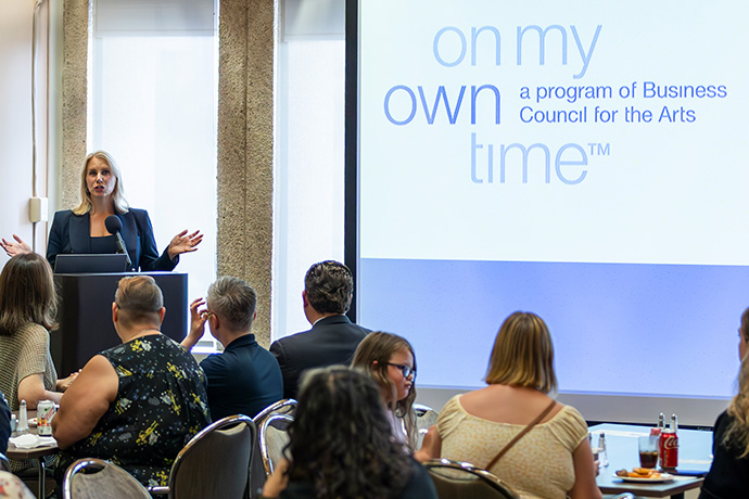 woman in blue suit with long blonde hair speaks from podium at left of a projected logo on screen in front of a seated audience at the On My Own Time 2024 art show