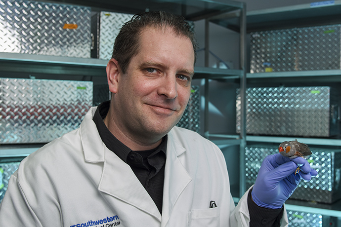 Smiling man with thin brown hair, wearing a white UT Southwestern lab coat, holding a small bird on his gloved hand.