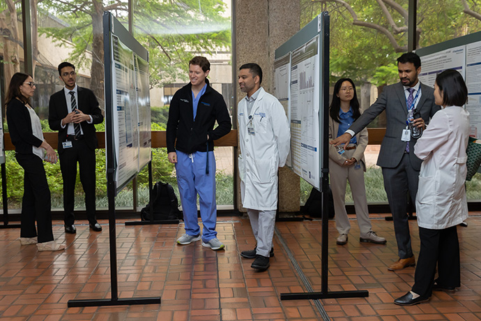 7 men and women in scrubs, white coats and suits in an atrium gather around boards displaying posters of current Internal Medicine research