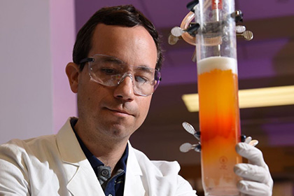 Man with dark hair and glasses staring intently at a beaker of orange liquid.