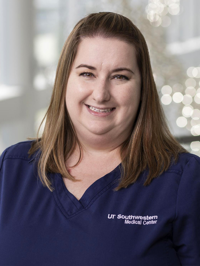Smiling woman with long brown hair, wearing blue UT Southwestern Medical Center scrubs.
