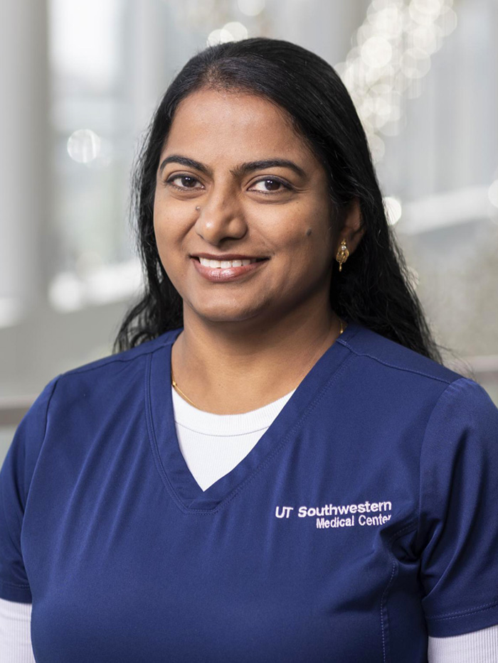 Smiling woman with long black hair, wearing blue UT Southwestern Medical Center scrubs over a white shirt.