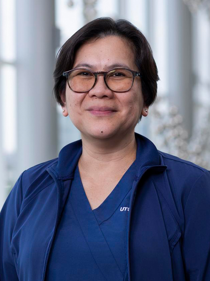 Smiling woman with short dark hair, wearing blue UTSW scrubs and jacket, earrings, and brown-rimmed glasses.