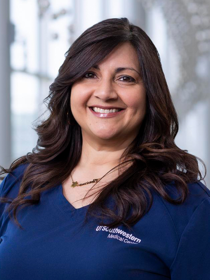 Smiling woman  with long dark curled hair, wearing blue UT Southwestern Medical Center scrubs and a necklace.