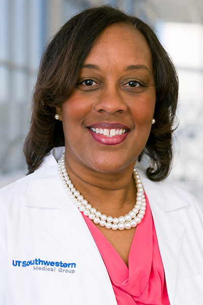 Smiling woman with dark hair wearing a white UT Southwestern Medical Center lab coat over a pink blouse, with a double strand of white pearls and pearl earrings.