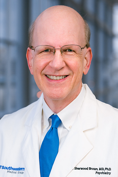 Smiling man with balding fair hair wearing a white UT Southwestern Medical Center lab coat, labeled with his name, Sherwood Brown, M.D., Ph.D., Psychiatry. He also wears a white shirt and blue tie and half-framed glasses.