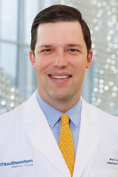 Smiling man with short dark hair wearing a white UT Southwestern Medical Center lab coat, labeled with his name, Brad Cutrell, M.D., Infectious Diseases, over a blue shirt and yellow tie.