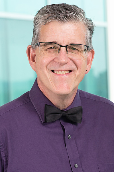 Smiling man with gray hair wearing a purple shirt, black bow tie, and half-rimmed glasses.