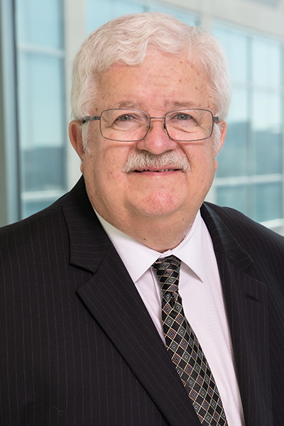 Smiling man with white hair and mustache, wearing a dark suit and tie with a white dress shirt and wire-framed rectangular glasses.